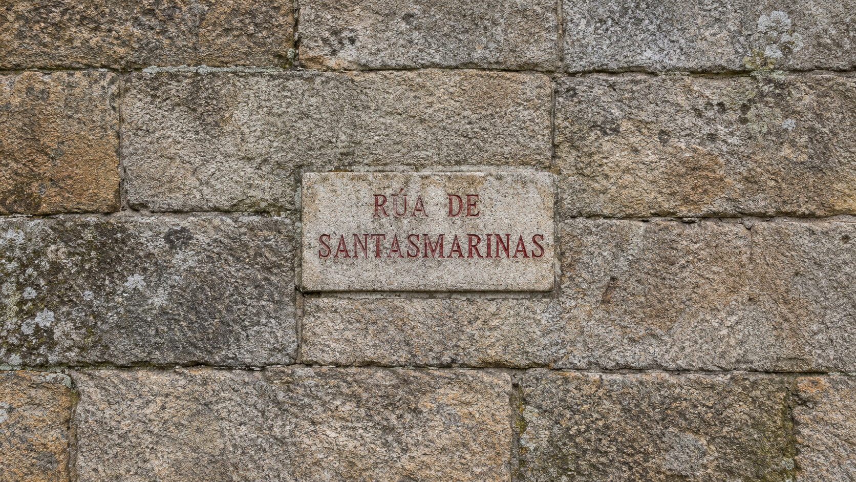 Placa de granito con letras vermellas: Rúa de Santasmarinas, sobre unha parede de pedra antiga de Santiago de Compostela.